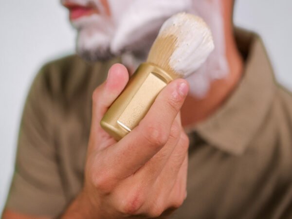 NATU synthetic shaving brush in gold held in the hand beside a tub of shaving cream