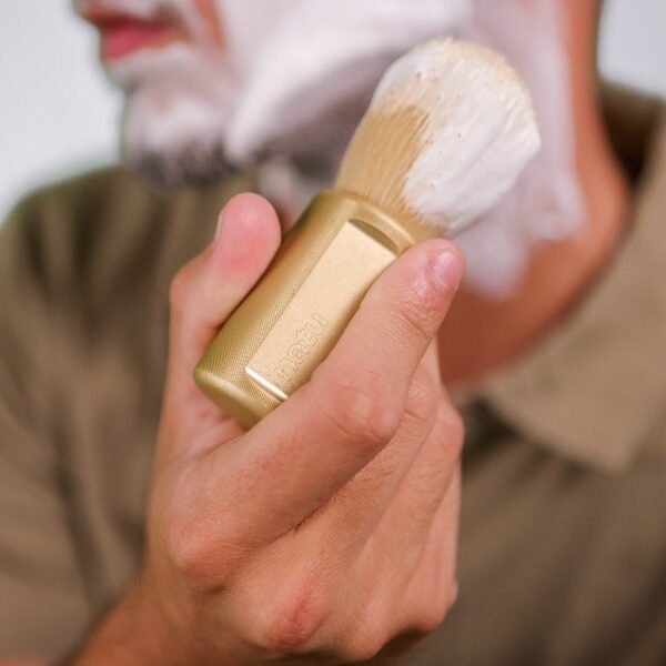 NATU synthetic shaving brush in gold held in the hand beside a tub of shaving cream