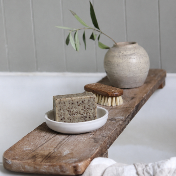 GROOM North Soap bar in a white dish on a wooden bath caddy beside a bristle brush